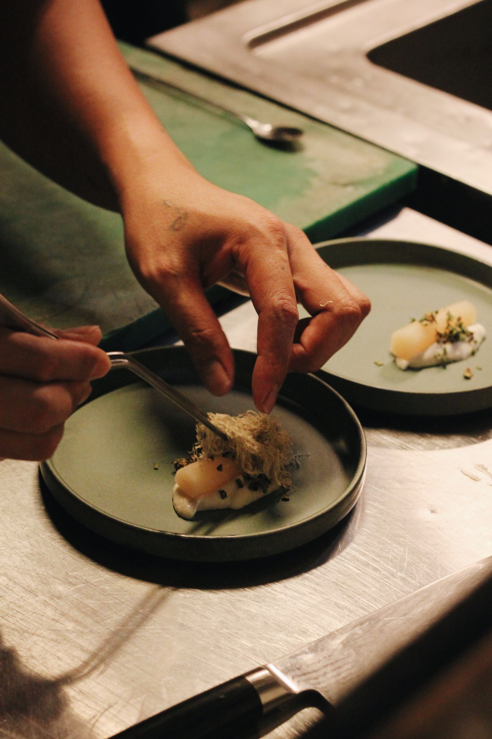 Hands plating a delicate course with tweezers in a warm, dim kitchen